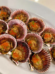 Close-up of baked scallop appetizers in purple ridged shells on a white plate, topped with crunchy breadcrumbs, orange fish roe and chopped chives
