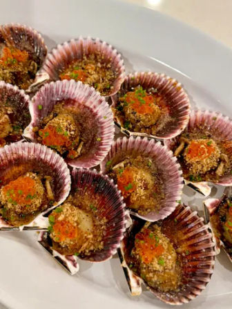 Close-up of baked scallop appetizers in purple ridged shells on a white plate, topped with crunchy breadcrumbs, orange fish roe and chopped chives