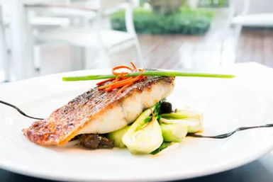 Pan-seared fish fillet with crispy skin atop sautéed bok choy and mushrooms, carrot curls and chive garnish on a white plate in a bright restaurant setting