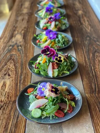 Row of plated spring salads with arugula, cherry tomatoes, cucumber slices and colorful edible pansy flowers on dark plates lined along a rustic wooden table — fresh farm-to-table salad presentation for a cafe or restaurant.