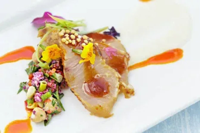 Fine-dining plate of thinly sliced sashimi-style fish drizzled with glossy sauce, garnished with edible flowers, microgreens, crunchy puffed grains and a colorful vegetable relish on a white plate.