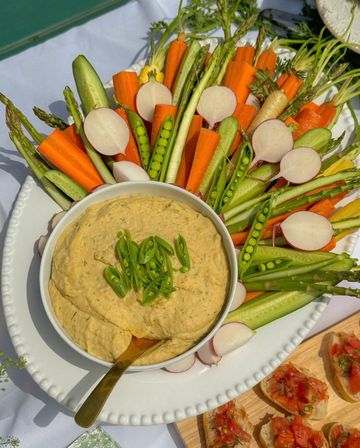 Creamy hummus garnished with sliced snap peas, surrounded by arranged fresh crudités—carrot sticks, radish rounds, asparagus, cucumber and sugar snap peas—on a white platter for a summer picnic.