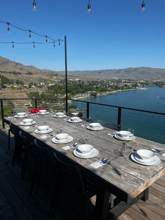Sunlit lakefront patio with a long rustic wooden dining table set for a large meal — white plates, bowls and wine glasses — string lights overhead and panoramic lake and mountain views.