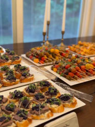 Vibrant catered appetizer spread on a buffet table: crostini topped with roast beef, caramelized onions and microgreens, alongside colorful cherry tomato and meat skewers on white platters.