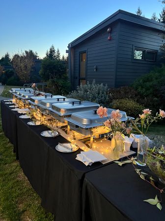 Outdoor evening buffet in a garden: row of silver chafing dishes on a black-clothed table lit by warm fairy lights, plates and napkins ready, pink floral arrangements, with a dark-blue cottage and trees in the background.