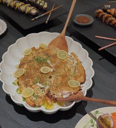 Lemon-herb pan-seared white fish fillets in a scalloped white serving bowl with wooden paddle, sushi rolls and dipping sauce on black slate in the background.