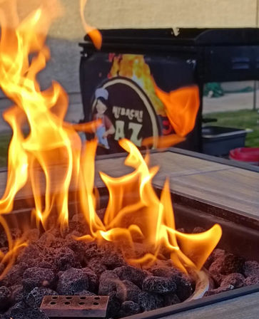 Close-up of a backyard patio fire pit with black lava rocks and dancing orange flames, blurred grill cart in the background.