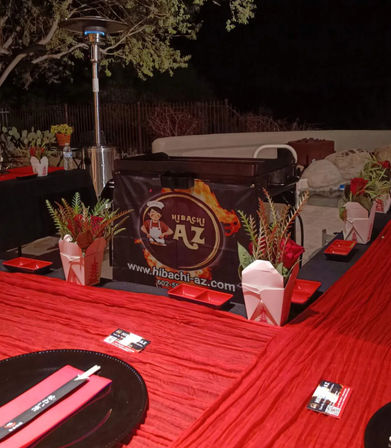 Nighttime outdoor hibachi setup on a red-draped backyard patio with a portable grill, patio heater, red takeout-box centerpieces and chopsticks.
