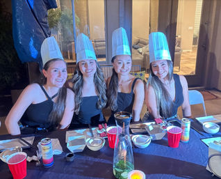 Four smiling women in tall paper chef hats at a backyard patio dinner party, seated at a table with plates, red cups, canned drinks, a carafe with lime slices and a candle.