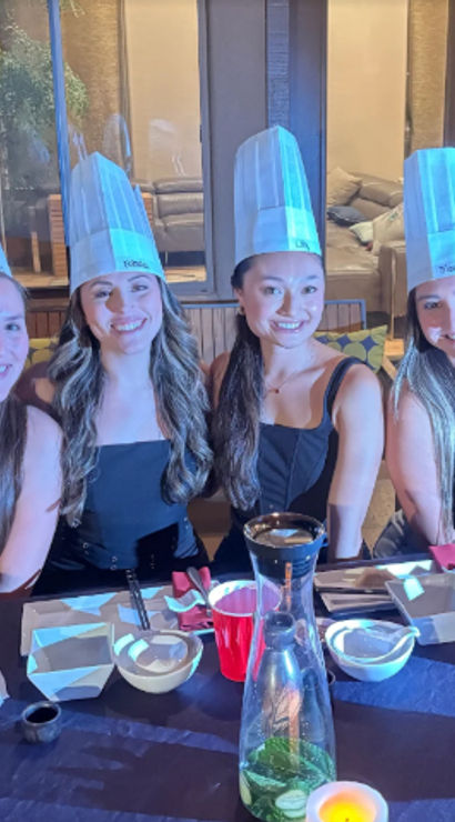 Four smiling women in tall paper chef hats at a backyard patio dinner party, seated at a table with plates, red cups, canned drinks, a carafe with lime slices and a candle.