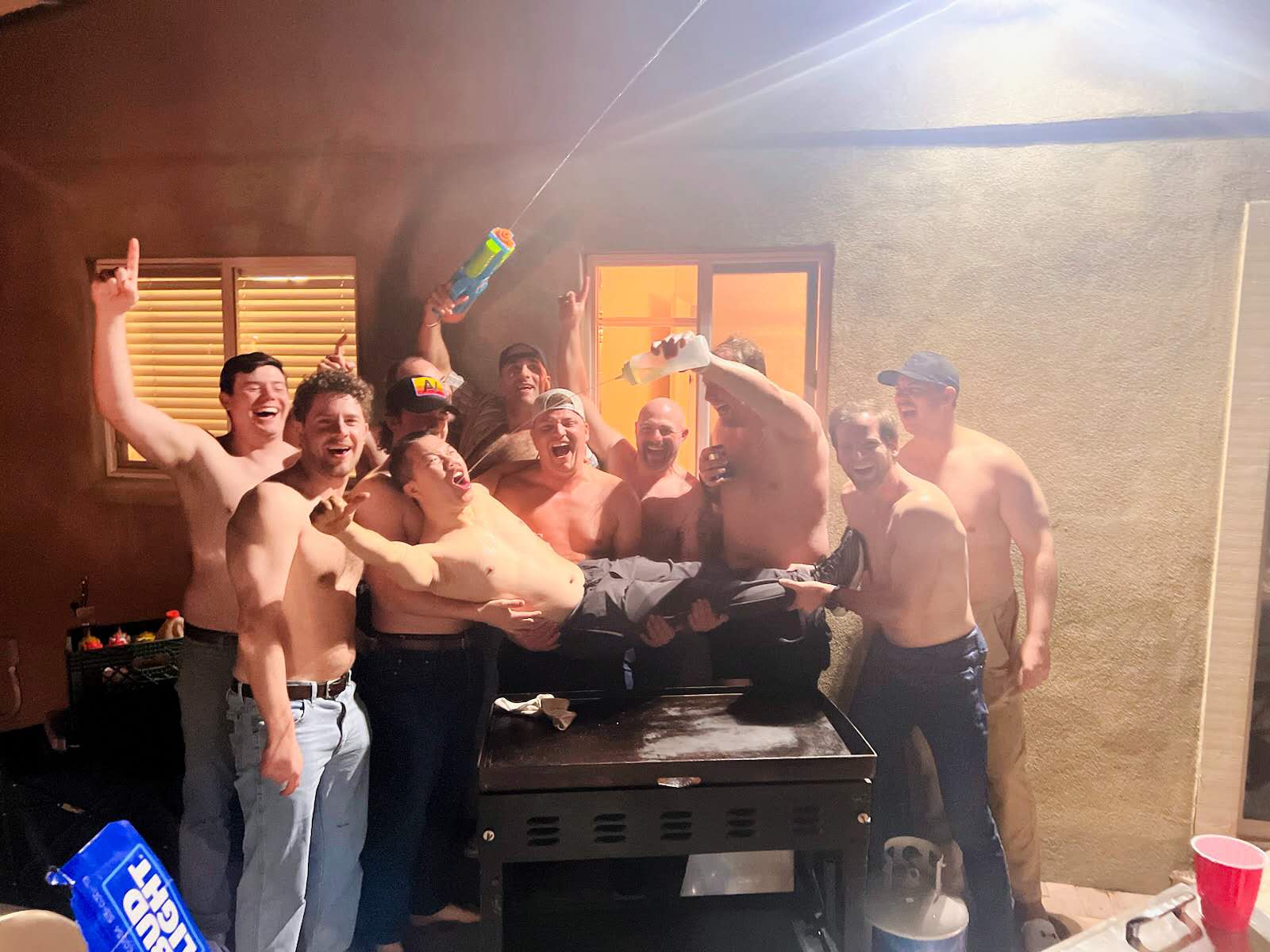 Group of shirtless men at a backyard patio party at night, hoisting a friend over a grill while others spray a water gun and pour drinks, with red solo cups and a propane tank visible.