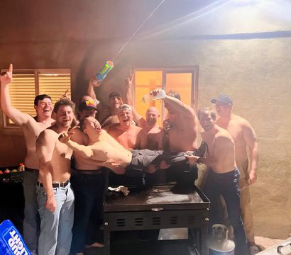Group of shirtless men at a backyard patio party at night, hoisting a friend over a grill while others spray a water gun and pour drinks, with red solo cups and a propane tank visible.