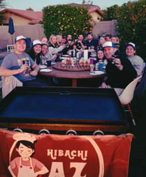 Cheerful backyard hibachi party: a large group of friends gathered around a round outdoor patio table, toasting with drinks behind a portable grill and chef banner.