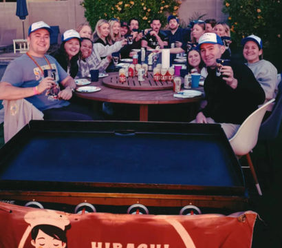 Cheerful backyard hibachi party: a large group of friends gathered around a round outdoor patio table, toasting with drinks behind a portable grill and chef banner.