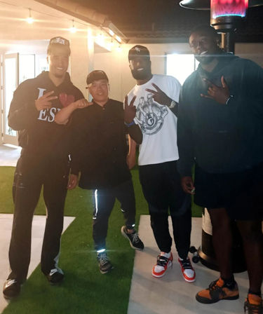 Group of four casually dressed people posing on a suburban backyard patio at night under string lights, standing on artificial turf near a tall outdoor heater, flashing peace and shaka hand signs and wearing streetwear sneakers.