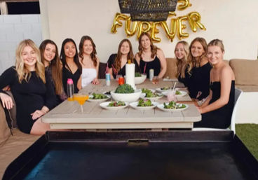 Nine friends gathered at an outdoor patio table for a casual celebration, plates of salad and drinks beneath gold "FUREVER" balloons.