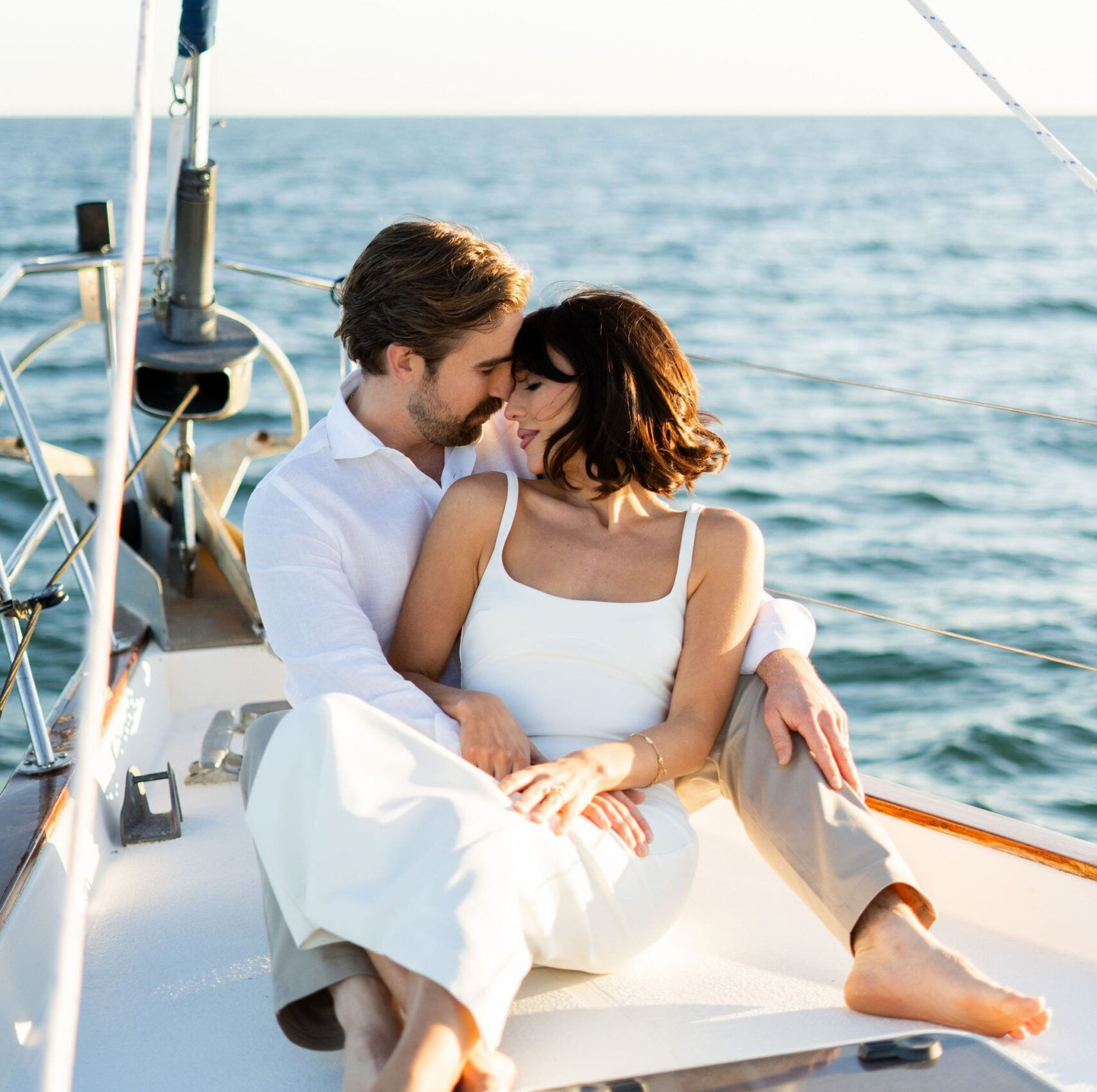 Romantic couple cuddling on the bow of a sailboat, enjoying calm blue ocean and warm golden sunlight