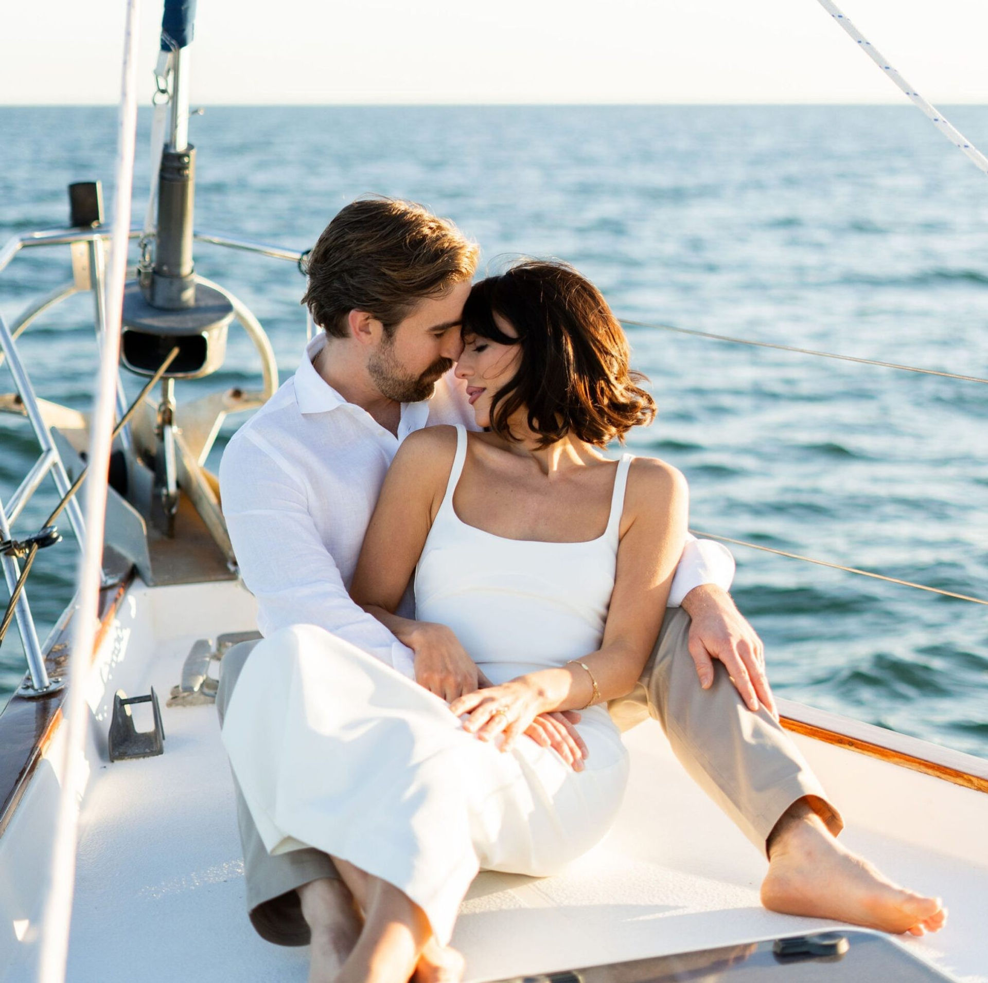 Romantic couple cuddling on the bow of a sailboat, enjoying calm blue ocean and warm golden sunlight