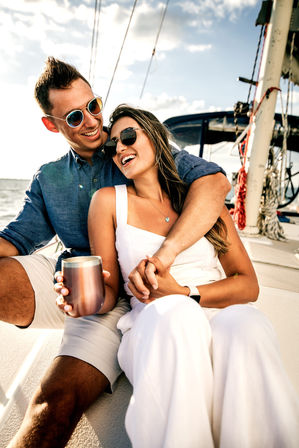 Smiling couple in sunglasses lounging on a sailboat deck, holding a travel tumbler and clasping hands against a sunny ocean backdrop
