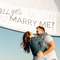 Romantic proposal on a sailboat — couple embracing under a white banner that reads 'MARRY ME?' against a clear blue sky