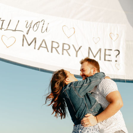 Romantic proposal on a sailboat — couple embracing under a white banner that reads 'MARRY ME?' against a clear blue sky