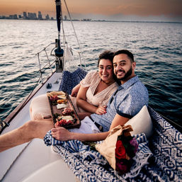 Smiling couple cuddling on the bow of a sailboat at sunset, sharing a charcuterie board and bouquet of roses with a distant urban skyline across calm coastal waters