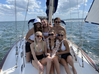Six friends in swimwear smiling on a sailboat deck under a blue sky, enjoying a sunny summer sailing trip on open coastal waters.