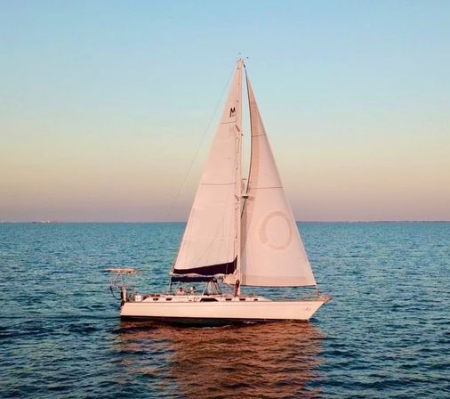 White sailboat cruising calm blue ocean near the horizon at golden sunset, tall mainsail and pastel sky reflecting on the water.