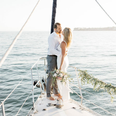 Romantic barefoot couple in wedding attire embracing on a sailboat bow, holding a floral bouquet above calm coastal waters at golden hour.