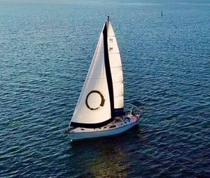 Aerial view of a single-masted white sailboat gliding across calm blue sea with a large circular emblem on its mainsail and sunlit ripples on the water