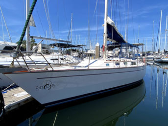 Sleek white sailboat with blue canvas cover docked in a sunny marina, tall masts and neighboring boats reflected in calm water.