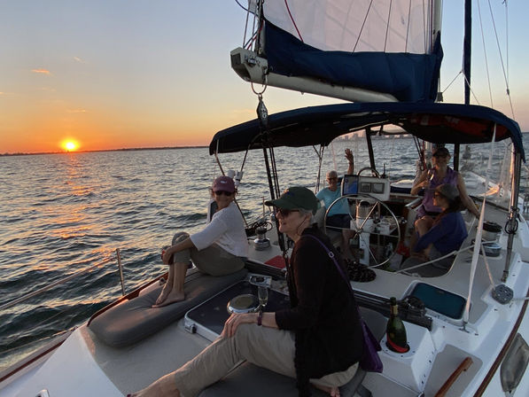 Cheerful group relaxing on a sailboat at sunset over calm coastal waters, passengers seated on deck and in the cockpit with a champagne bottle nearby and the orange sun low on the horizon.