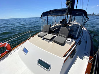 Sailboat deck and cockpit with two gray lounge cushions and a life ring, cruising on calm blue water under a clear sky with a distant coastal skyline.