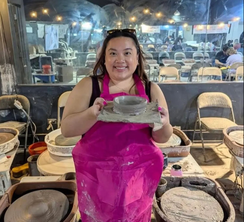Smiling person in a bright pink apron at a pottery wheel holding a freshly thrown clay bowl in a busy ceramics studio.