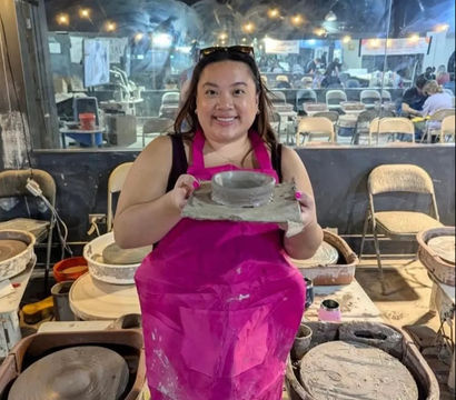 Smiling person in a bright pink apron at a pottery wheel holding a freshly thrown clay bowl in a busy ceramics studio.