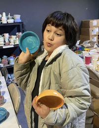 Young person playfully holding turquoise and orange ceramic bowls in a pottery workshop, surrounded by shelves of handmade ceramics and craft supplies.