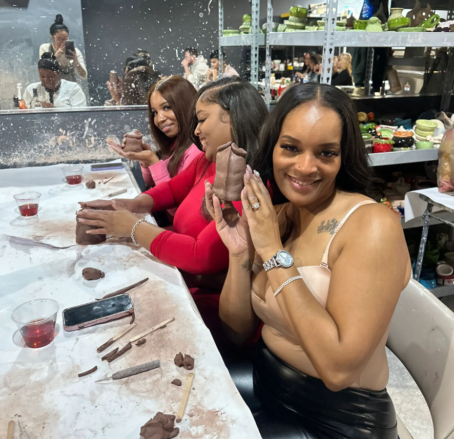 Three smiling women at a pottery studio shaping hand-built clay pieces during a group ceramics class, with sculpting tools, drinks on the table, and shelves of finished pottery in the background.