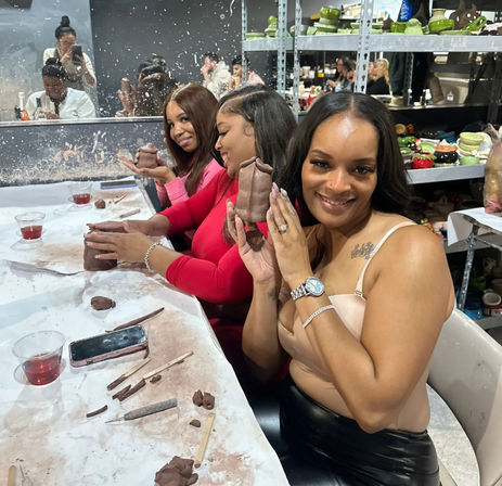 Three smiling women at a pottery studio shaping hand-built clay pieces during a group ceramics class, with sculpting tools, drinks on the table, and shelves of finished pottery in the background.