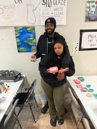 Grinning duo posing inside a pottery-painting studio, holding bright pink and teal small molds and ceramic pieces at a craft table with paints and a lily-pad canvas on the wall.