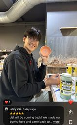 Smiling person in glasses and a black hoodie holds a small pink hand-painted ceramic bowl decorated with a yellow flower in a pottery studio at a workbench with glaze jars and clay tools.