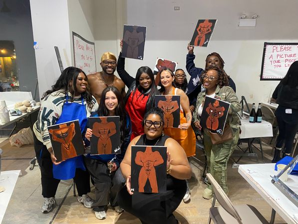 Smiling group in an indoor paint-and-sip art class holding canvases of stylized male torso paintings, wearing aprons and casual clothes in a lively studio setting with tables, easels and drinks.