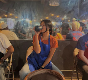 Participant in a blue apron sipping from a mug while seated at a pottery wheel in a lively indoor ceramics workshop with string lights.