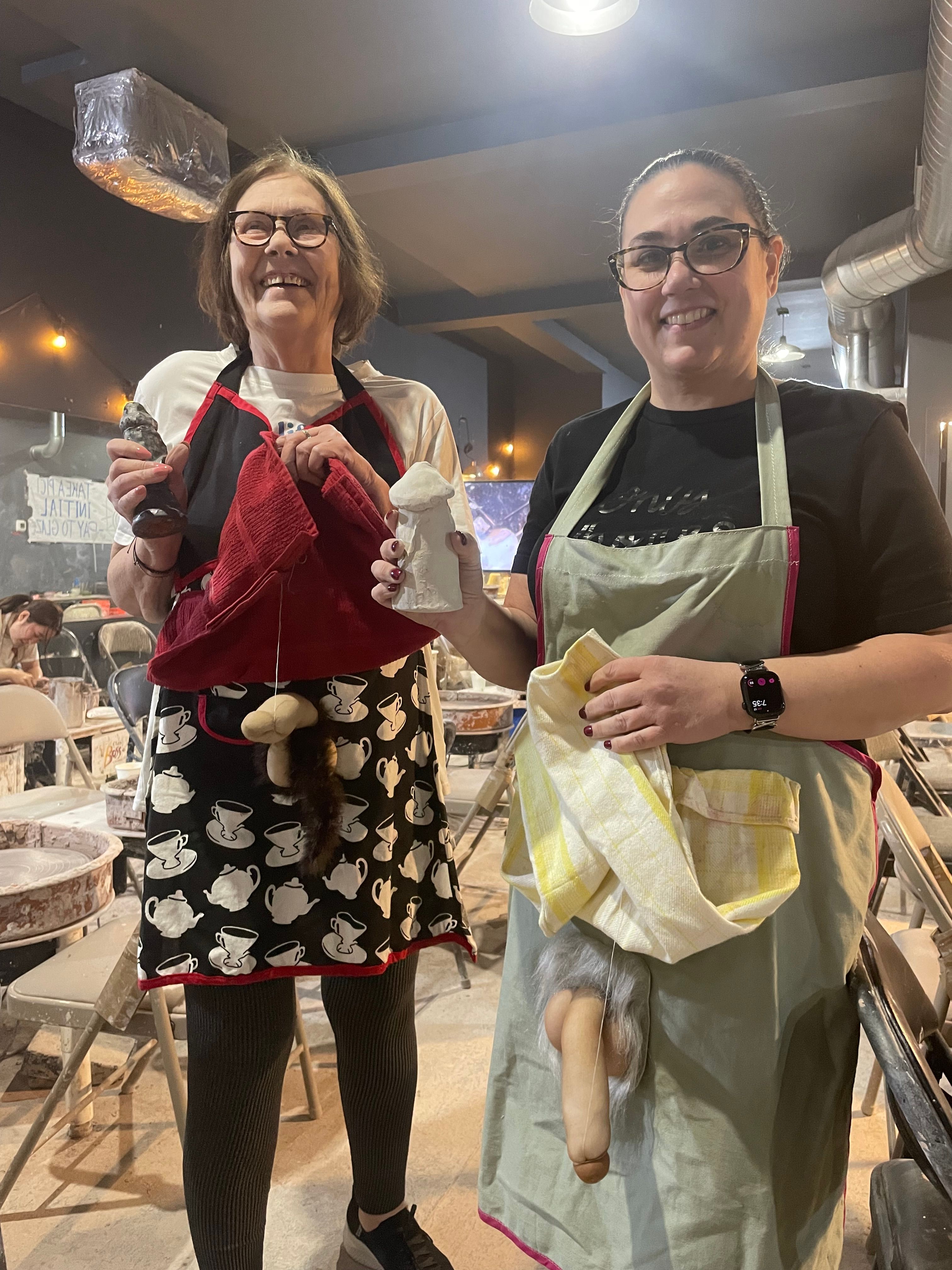 Two smiling people in a pottery studio wearing aprons, holding handmade clay sculptures and playful novelty phallic pieces — ceramics class setting