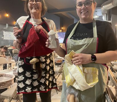 Two smiling people in a pottery studio wearing aprons, holding handmade clay sculptures and playful novelty phallic pieces — ceramics class setting