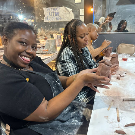 Smiling woman proudly holds a hand-sculpted clay vase during a lively indoor pottery class, friends shaping clay at a messy table with aprons, tools and a mirror-reflected studio background.
