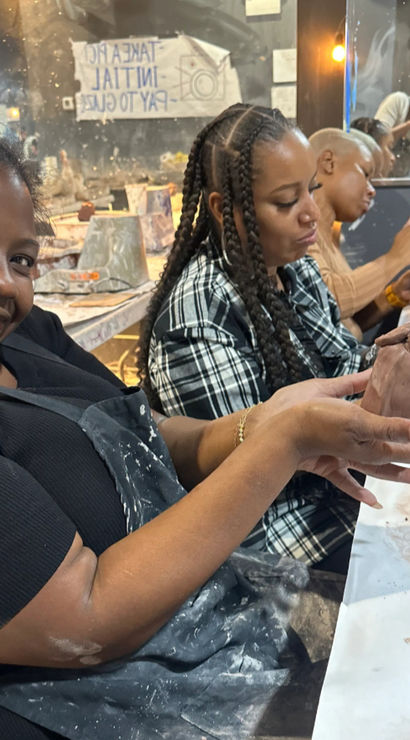 Smiling woman proudly holds a hand-sculpted clay vase during a lively indoor pottery class, friends shaping clay at a messy table with aprons, tools and a mirror-reflected studio background.