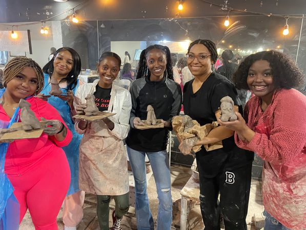 Six women in aprons smiling and holding small handmade clay sculptures on boards inside a cozy pottery workshop with string lights and mirrored wall.
