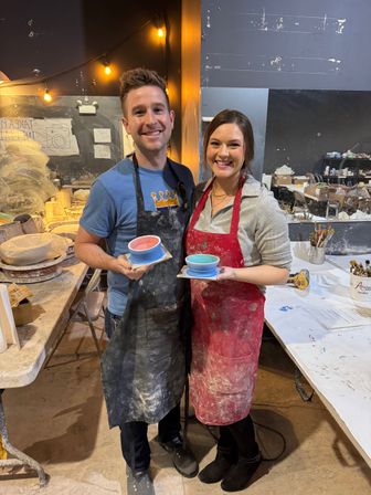 Two smiling people in paint-splattered aprons proudly holding handmade glazed ceramic bowls in a lively indoor pottery studio workshop with worktables, brushes, and string lights