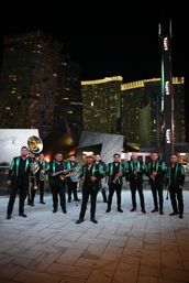 Brass band in matching green sequined jackets posing with trombones, trumpets, sousaphone and drums on a tiled plaza at night with Las Vegas high-rise hotels and illuminated skyline behind them.