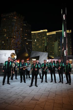 Brass band in matching green sequined jackets posing with trombones, trumpets, sousaphone and drums on a tiled plaza at night with Las Vegas high-rise hotels and illuminated skyline behind them.